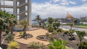 A modern building with multiple floors, a landscaped area with palm trees, and a distinctive triangular structure in the background. - Vacation Studio 'Primera Linea' with Sea View, Balcony and Wi-Fi (Las Palmas de Gran Canaria)