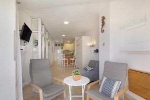 A modern living room with a sofa, chairs, a small table with a plant, a TV mounted on the wall, and a kitchen in the background. - Vacation Studio 'Primera Linea' with Sea View, Balcony and Wi-Fi (Las Palmas de Gran Canaria)