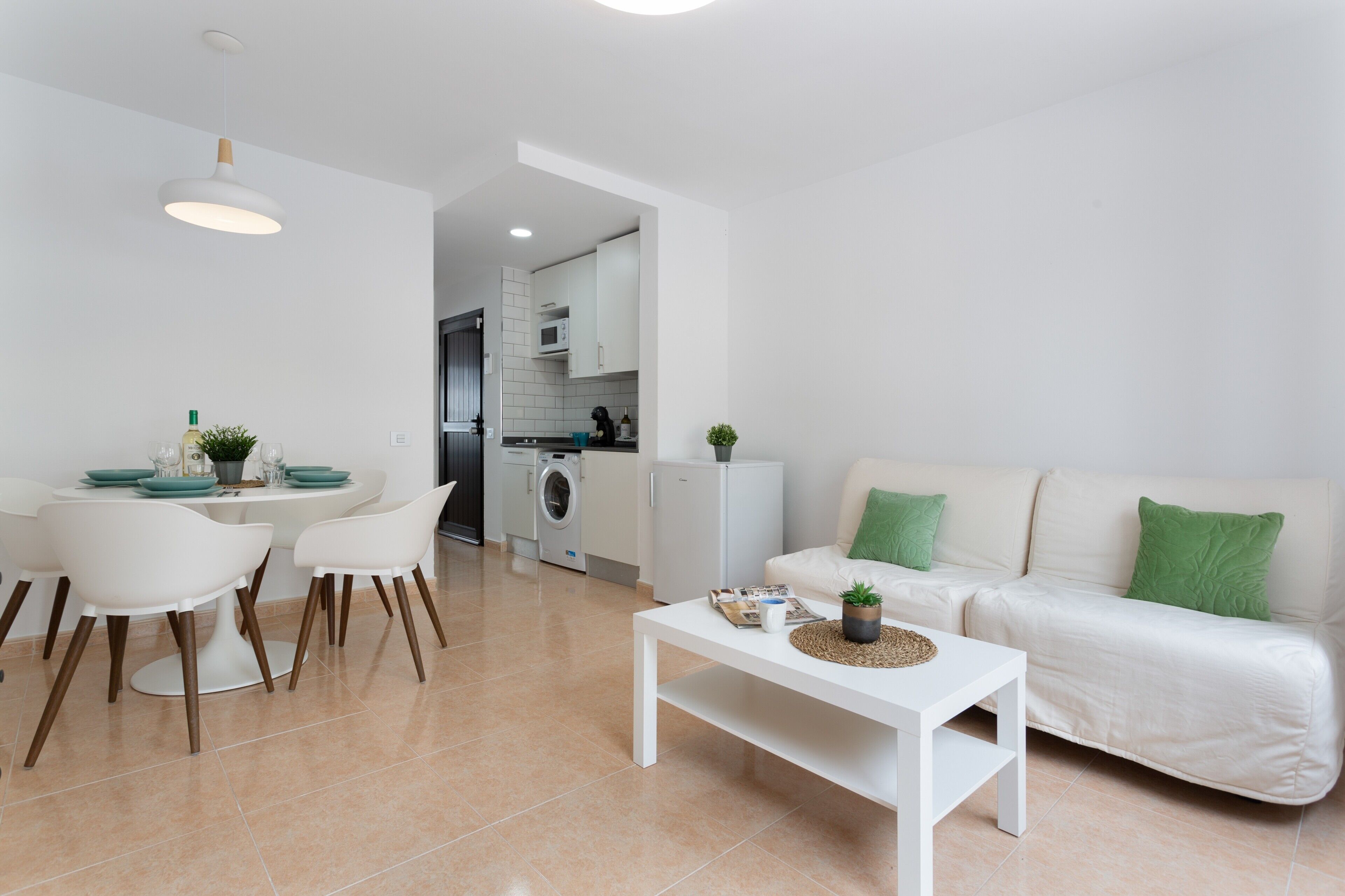 A modern living room with a white sofa, a dining table with chairs, a coffee table, and a kitchen area in the background.