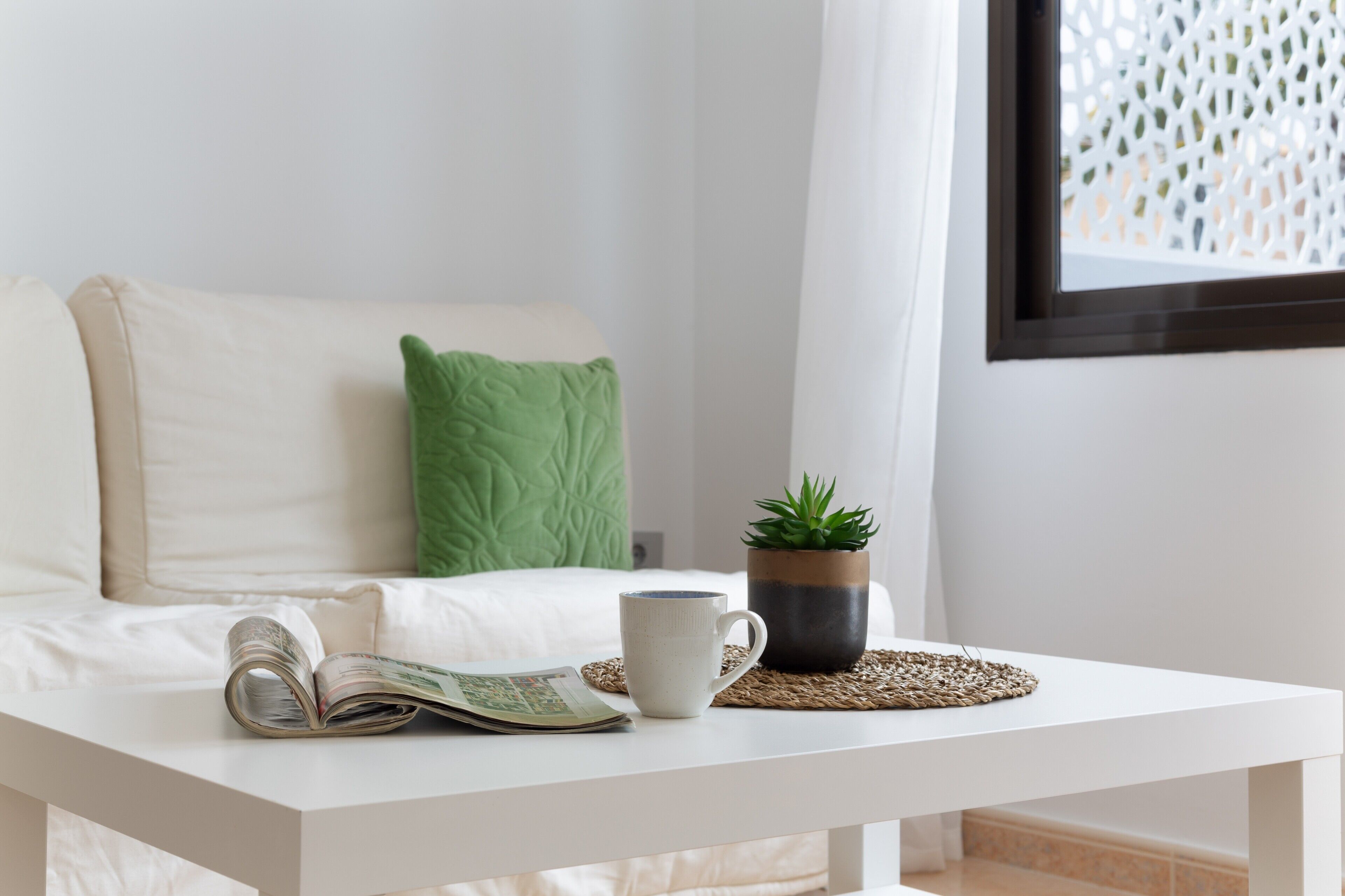 A white sofa with a green pillow, a white coffee table with a magazine, a white mug, and a small potted plant.