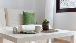 A white sofa with a green pillow, a white coffee table with a magazine, a white mug, and a small potted plant. - 'VV Oasis Duna Punto Alegre' Vacation with Shared Pool, Private Terrace, and Wi-Fi (Corralejo)
