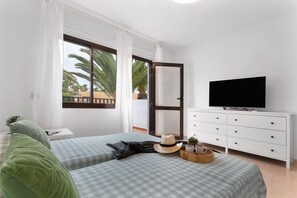 Ein Hotelzimmer mit zwei Betten, einem Fernseher, einem Schminktisch und einem Balkon mit Blick auf Palmen.