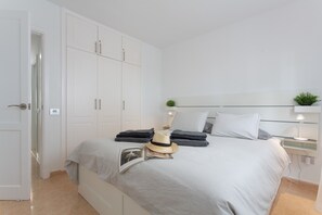 Un dormitorio con cama, armario, mesita de noche con lámpara y una planta pequeña.