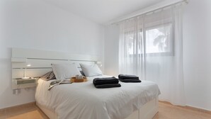 A neatly made bed with pillows and folded towels, a bedside table with a lamp, and a window with sheer curtains. - 'VV Oasis Duna Punto Alegre' Vacation with Shared Pool, Private Terrace, and Wi-Fi (Corralejo)
