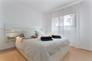 Una cama bien hecha con almohadas y toallas dobladas, una mesita de noche con lámpara, y una ventana con cortinas translúcidas.