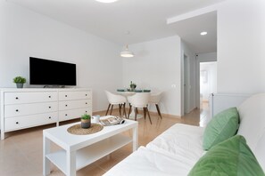 Una sala de estar moderna con un televisor de pantalla plana, un sofá blanco, una mesa de comedor con sillas y un armario blanco.