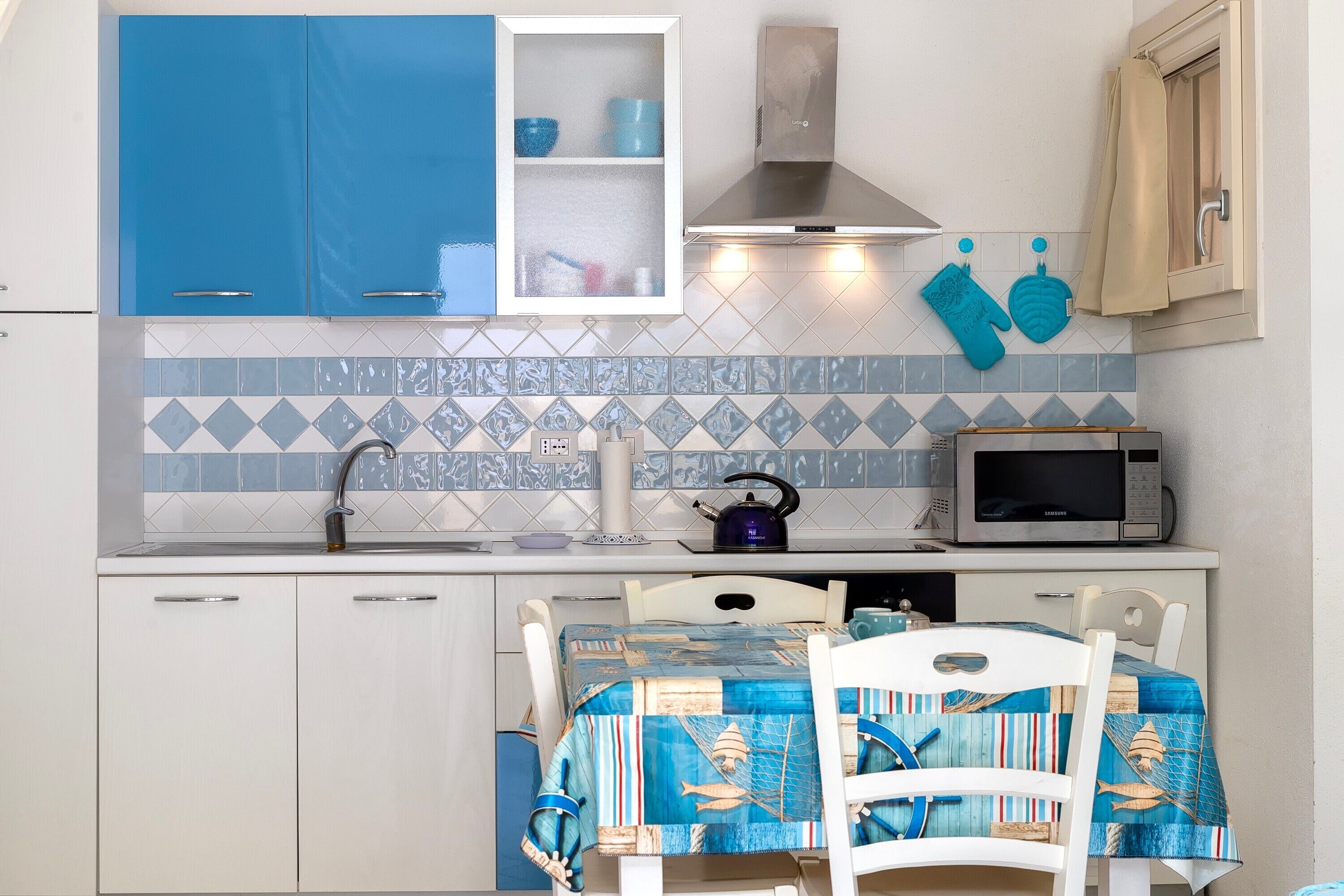 A compact kitchen with blue cabinets, a microwave, a kettle, and a table with a blue tablecloth.
