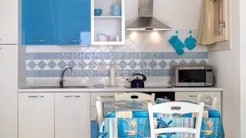 A compact kitchen with blue cabinets, a microwave, a kettle, and a table with a blue tablecloth.