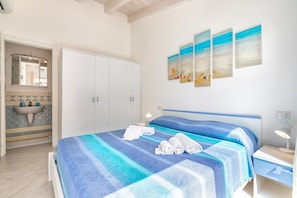 A bedroom with a bed, a nightstand, a lamp, a bathroom with a sink and mirror, and artwork on the wall.