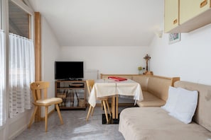 Une pièce avec un canapé beige, une chaise en bois, une petite table recouverte d’une nappe blanche, un téléviseur et un radiateur.