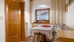 Ein Zimmer mit Holztür, Tisch mit weißem Tuch, Stuhl, Bank, Fenster mit Blick ins Grüne und einer Hängeleuchte.