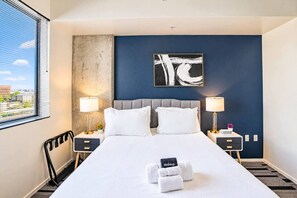 A hotel room with a large bed, two bedside tables, a headboard, a window with blinds, and a city view.
