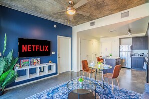 A modern living room with a large TV displaying "NETFLIX," a dining area with a glass table, and a kitchen in the background.