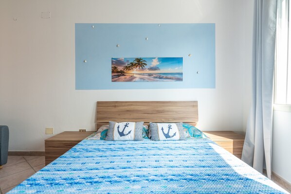 A bedroom with a bed, a painting of a beach scene, and a window with curtains.