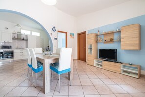 Une cuisine moderne avec des meubles blancs, un coin repas avec une table et des chaises en bois, un téléviseur posé sur un meuble en bois et un mur bleu.