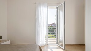 Une chambre à coucher avec un lit, une fenêtre avec des rideaux et une vue sur un balcon et des bâtiments à l’extérieur.