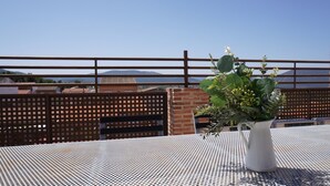 Un jarrón blanco con follaje verde sobre una mesa metálica con superficie perforada, frente a una valla de madera que da a edificios y montañas al fondo.