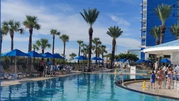 A swimming pool area with palm trees, blue umbrellas, and lounge chairs.