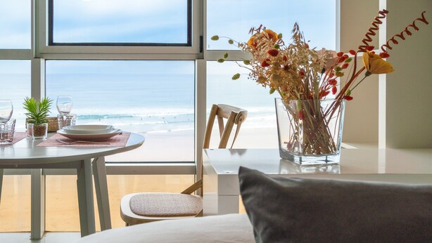 Une chambre avec vue sur la plage, une table à manger avec des assiettes et des verres, un vase rempli de fleurs et un lit avec un oreiller.