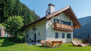A chalet with a balcony, a dining area with benches, and a view of a forested mountain.