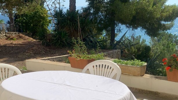 Un área de estar al aire libre con una mesa redonda y sillas, rodeada de plantas en macetas y vista a un cuerpo de agua.