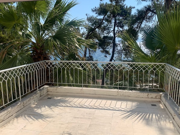 A balcony with a white metal railing, a tiled floor, and a view of palm trees and pine trees.