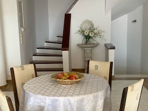 Un coin repas comprenant une table ronde, des chaises, un panier de fruits, un escalier et un élément décoratif mural.