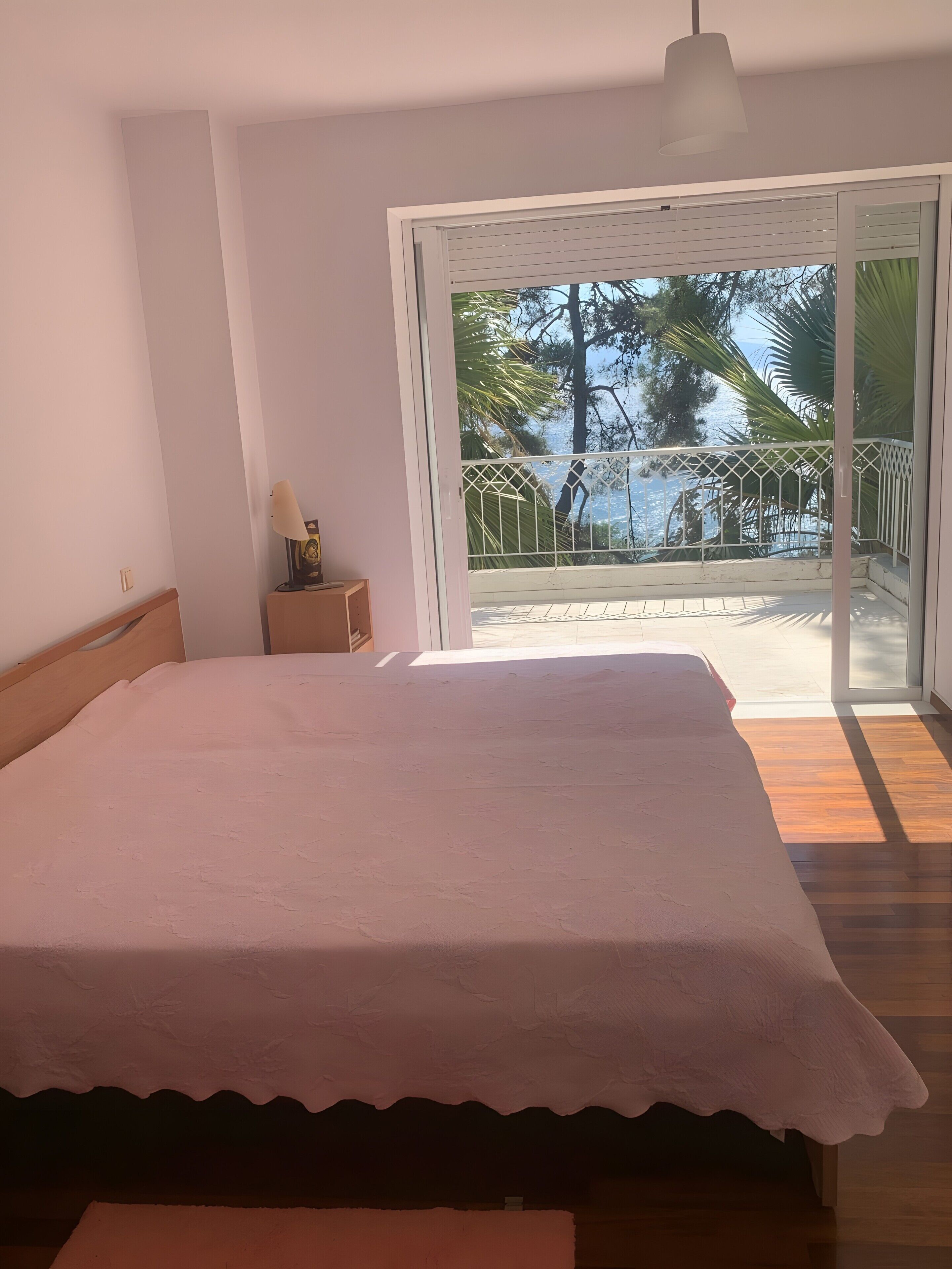 A bedroom with a bed, a nightstand, a lamp, and a view of a balcony with a railing.