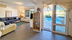 A living room with a sofa, a coffee table, a TV, and a bookshelf. There is a sliding glass door leading to a courtyard. - Villa 'Beach , Swimming Pool For 8' with Sea View, Wi-Fi and Air Conditioning (Thermisia)