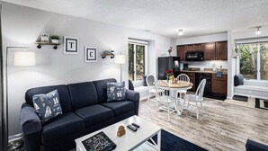 Un salon moderne avec un canapé bleu foncé, des chaises blanches, une table basse et un coin cuisine doté de meubles en bois et d’un four à micro-ondes.