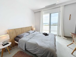 Une chambre d’hôtel avec un lit, une petite table avec une lampe, une chaise et une vue sur la mer par une grande fenêtre.