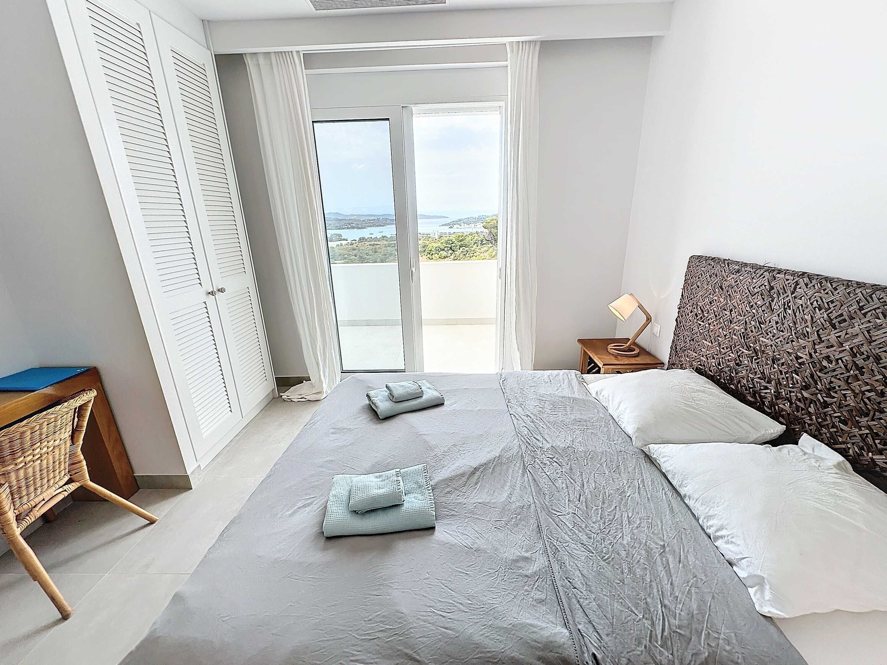 Un dormitorio con cama, mesita de noche, lámpara, una silla de mimbre y vistas a través de un ventanal.