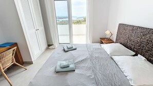 Et soveværelse med seng, et natbord, en lampe, en fletstol og udsigt gennem en glasdør.