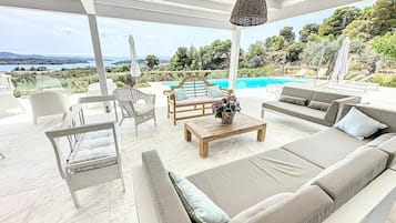 Un espacio de estar techado al aire libre con piscina, mobiliario blanco y vistas al paisaje.