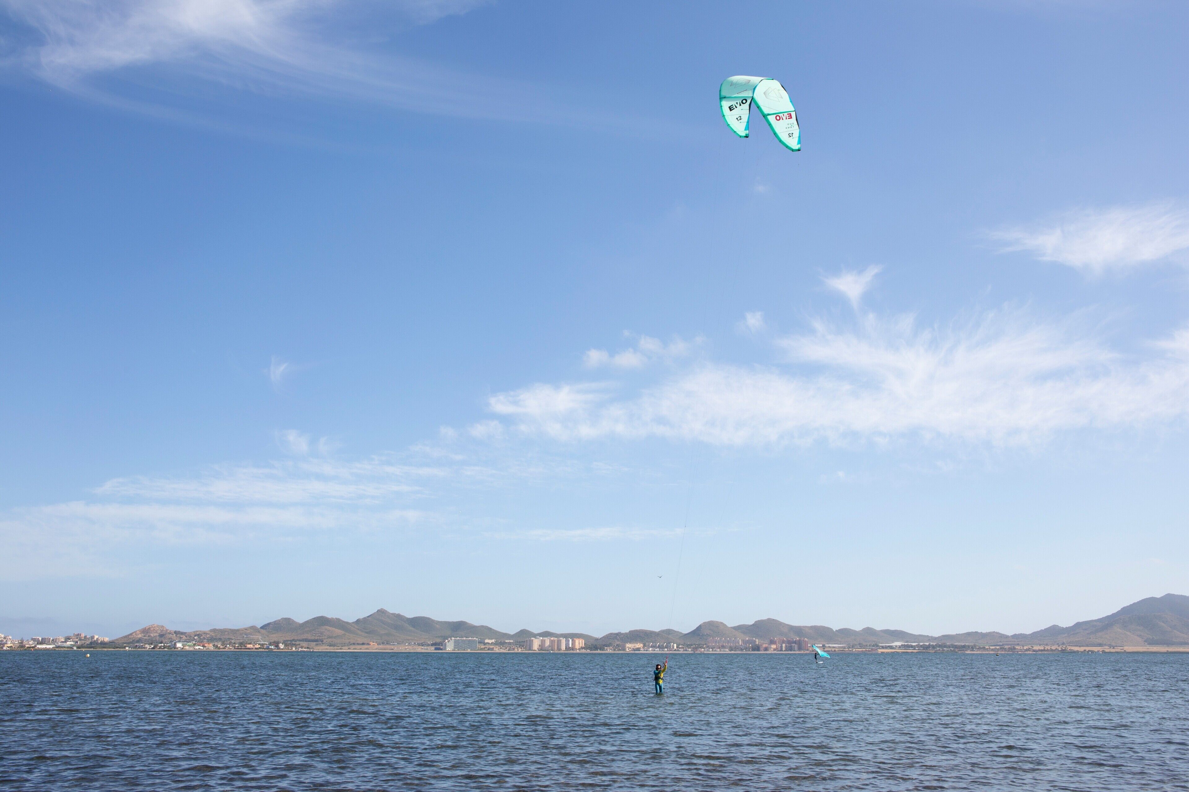 A coastal view with a person kiteboarding on the water and mountains in the background.