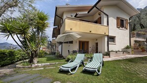 A two-story house with a balcony, a garden with a palm tree, and two lounge chairs. - Apartment 'Edda - Sole' with Lake View, Private Terrace, and Wi-Fi (Brenzone sul Garda)
