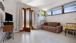 A living room with a sofa, a table with a floral tablecloth, a television on a stand, and a window with curtains. - Apartment 'Edda - Sole' with Lake View, Private Terrace, and Wi-Fi (Brenzone sul Garda)