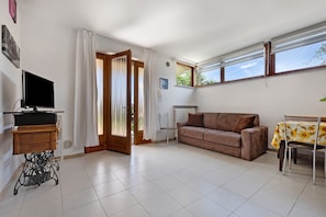 A living room with a sofa, a table with a floral tablecloth, a television on a stand, and a window with curtains.