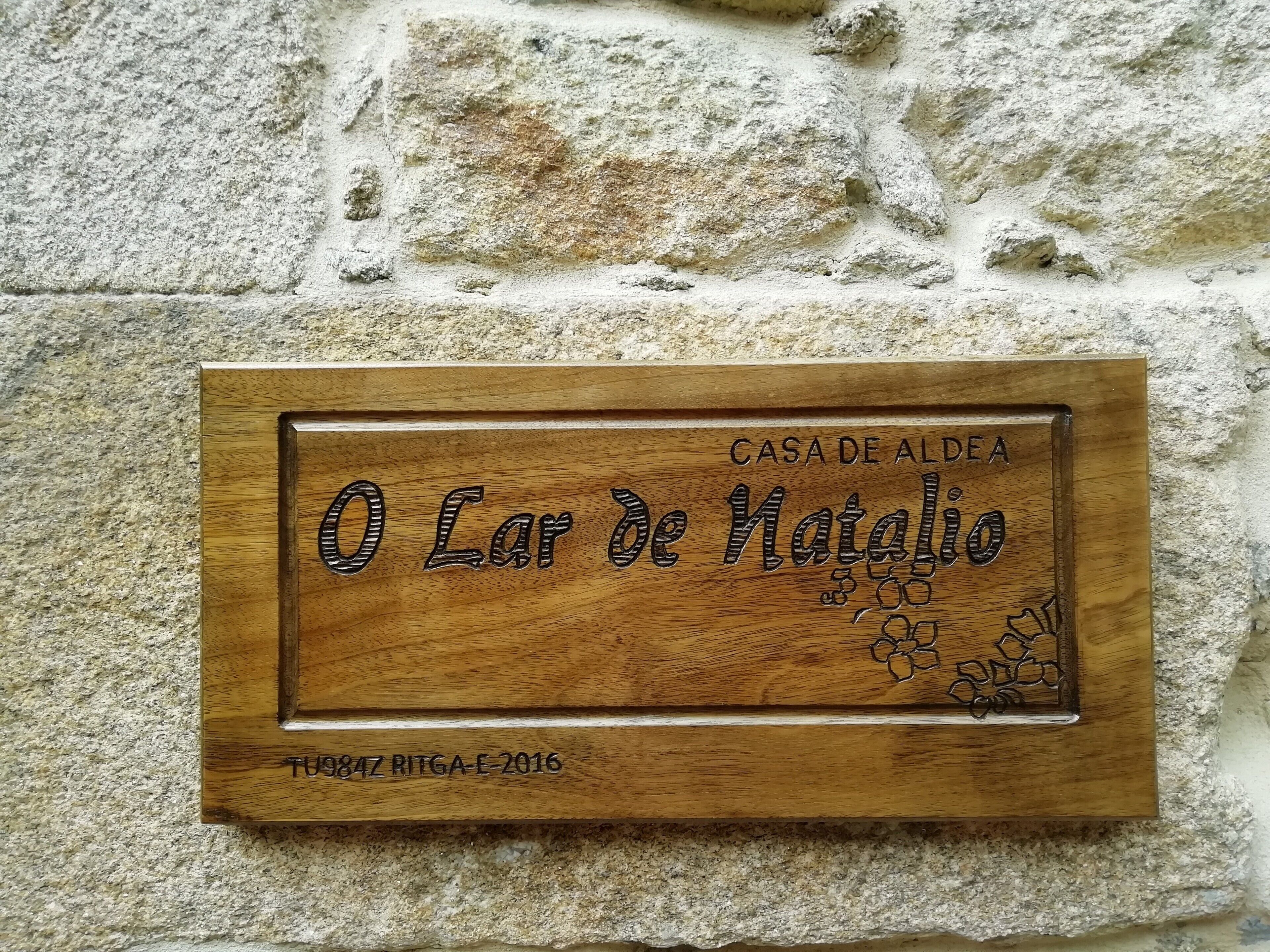 Eine Holztafel mit der eingravierten Aufschrift 'CASA DE ALDEA' und 'O Lar de Natalio', die an einer Steinsäule angebracht ist.