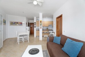 Een woonkamer met een bruine bank, een eettafel met stoelen, een keuken op de achtergrond en een plafondventilator.