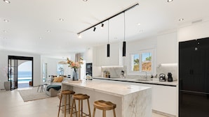 Una cocina moderna con una isla de mármol, taburetes de madera, luces colgantes y vistas al exterior.