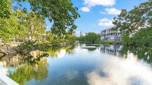 Une rivière paisible avec un quai, bordée d’une végétation luxuriante et surplombée d’un immeuble doté de nombreux balcons.