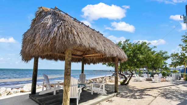 Une terrasse en bois située au bord de la plage, dotée d’un toit de chaume, de chaises blanches et offrant une vue sur l’océan.