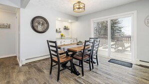 A dining area with a wooden table and chairs, a clock on the wall, and a view through large windows.
