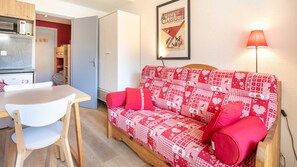 A compact living space with a kitchenette, a dining table, a sofa with red cushions, and a framed poster on the wall.