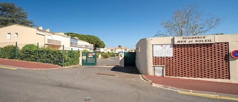 Un quartier résidentiel doté d’une entrée sécurisée par un portail, d’une enseigne indiquant « RÉSIDENCE MER ET SOLEIL » et d’un panneau interdisant le stationnement.