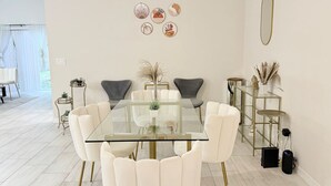 A modern dining room with a glass table surrounded by white chairs, a gold-framed side table, and decorative wall art. - 3BR | Spacious | Entire House | Close to all Parks (Orlando)