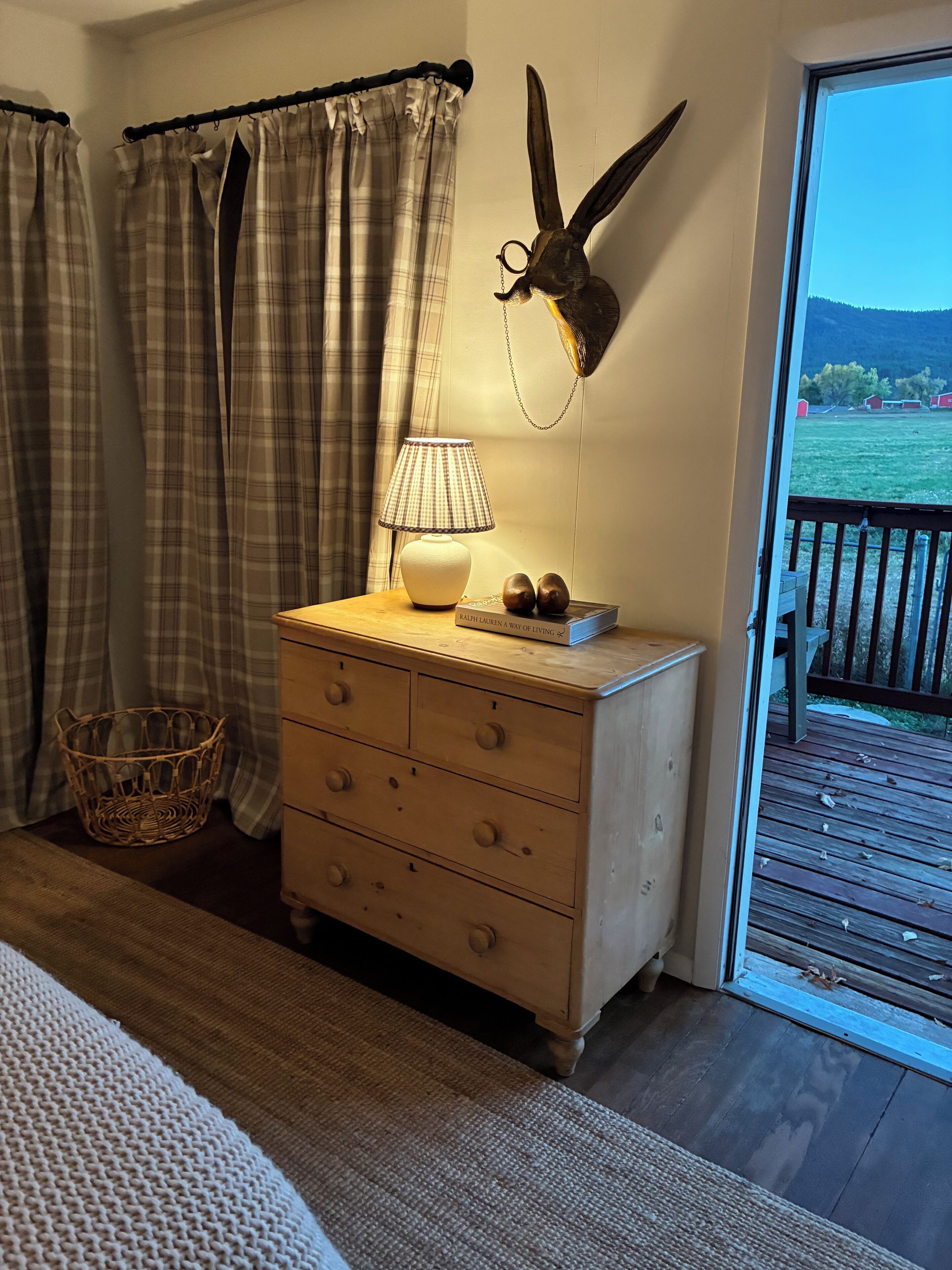 Ein hölzerner Kommode mit einer Lampe, einer Wanddekoration mit einem Hasen, Karottenvorhängen und Blick auf eine Holzterrasse und viel Grün draußen.
