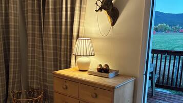Ein hölzerner Kommode mit einer Lampe, einer Wanddekoration mit einem Hasen, Karottenvorhängen und Blick auf eine Holzterrasse und viel Grün draußen.