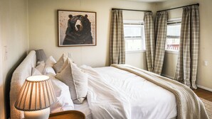 Ein Schlafzimmer mit Bett, Kissen, einer Lampe, einem Tablett, einem Bärenbild und karierten Vorhängen.